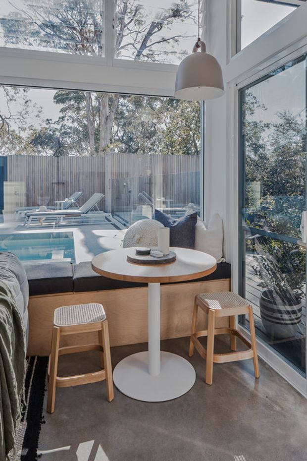 Bright, compact sunroom or indoor-outdoor lounge with extensive white-framed glazing overlooking a small swimming pool and fenced courtyard. The space features a built-in timber bench with dark cushions, a round white pedestal table, woven timber stools, a large neutral pendant light, polished concrete flooring, and a potted plant. Natural daylight and strong indoor-outdoor connection dominate the scene.