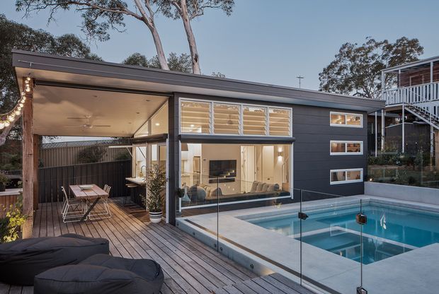 A contemporary single-storey home viewed from an elevated timber deck, with dark horizontal cladding, broad glazing, a covered outdoor dining and lounge area, and a swimming pool enclosed by frameless glass balustrades. Warm interior lighting is visible through the windows, while mature trees and a neighbouring raised structure surround the property.