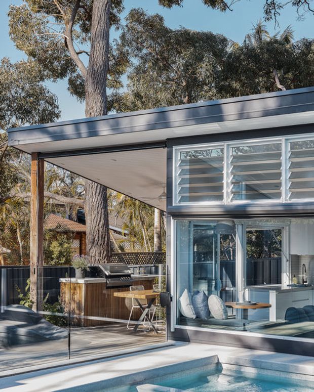 A contemporary residential outdoor entertaining area beside a swimming pool, viewed toward a glazed living space. The scene features a flat-roofed extension with dark cladding, extensive aluminium-framed glazing and louvred windows, a covered timber deck, built-in outdoor kitchen with barbecue, dining furniture, and a glass pool barrier. Mature trees surround the home.
