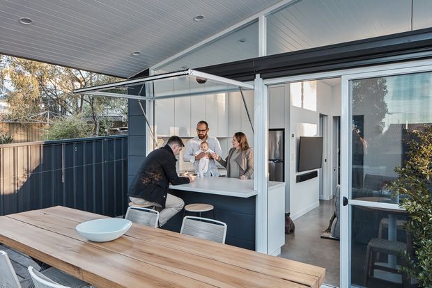 A contemporary covered outdoor entertaining area with a built-in outdoor kitchen or bar beneath a retractable awning. A family gathers around the counter, while a large timber dining table occupies the foreground. Dark blue-grey cabinetry and screening contrast with white framed glazing, a pale panelled ceiling, and a sliding glass door leading into the bright interior.