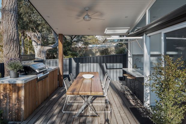 A covered outdoor dining and entertaining deck beside a glazed home extension. The space features a timber dining table with white metal-framed chairs, a built-in barbecue set into timber cabinetry, a ceiling fan, overhead heater or skylight elements, timber decking, and dark vertical privacy screening. Large windows and a servery-style opening connect the deck to the interior, with mature trees visible beyond.