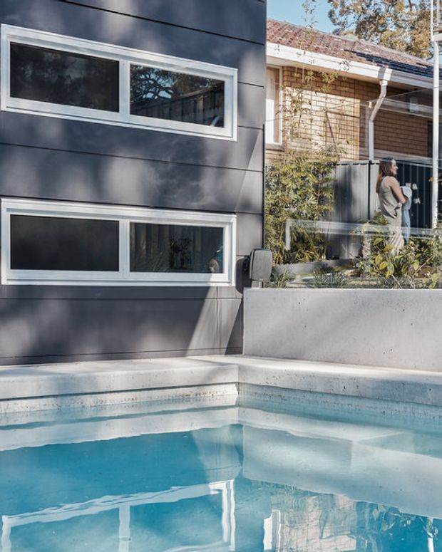 Exterior view of a residential swimming pool beside a contemporary dark-clad building. The scene shows horizontal white-framed windows, a smooth concrete planter wall, pale pool coping, and a neighbouring brick structure with a tiled roof and screened garden. Blue pool water and reflections dominate the foreground.