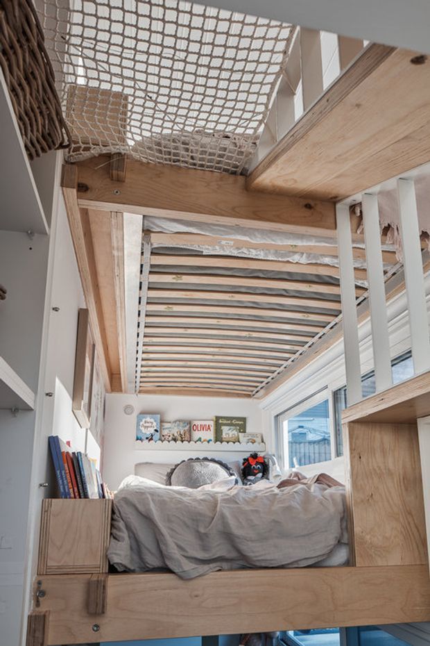 A compact loft-style bedroom viewed from a lower level, featuring a raised timber sleeping platform with exposed plywood and timber framing. A white rope safety net spans the open loft edge above, while the bed sits beneath a partially open slatted ceiling structure. Built-in timber shelving and storage, books, soft bedding, and children’s decorative items create a playful, natural interior. Large windows bring in daylight along the right side.