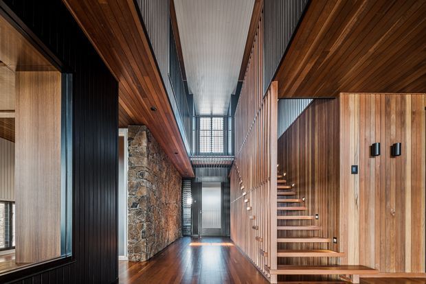 A dramatic contemporary interior hallway with a timber-lined ceiling and polished dark timber floor. The space features a sculptural open staircase integrated into vertical timber wall panelling, a rugged exposed stone feature wall, dark ribbed wall surfaces, and a tall glazed clerestory-like void that brings in natural light. The composition emphasizes strong linear geometry, natural materials, and a warm yet refined atmosphere.