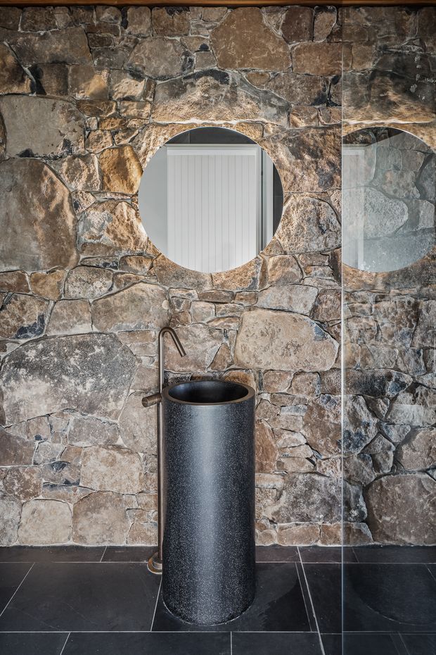 Compact bathroom interior featuring a freestanding cylindrical black basin with matching dark wall-mounted tap, set against a rustic irregular natural-stone wall. A round mirror and partial glass shower screen are visible, with large dark floor tiles creating a dramatic, textural scheme.