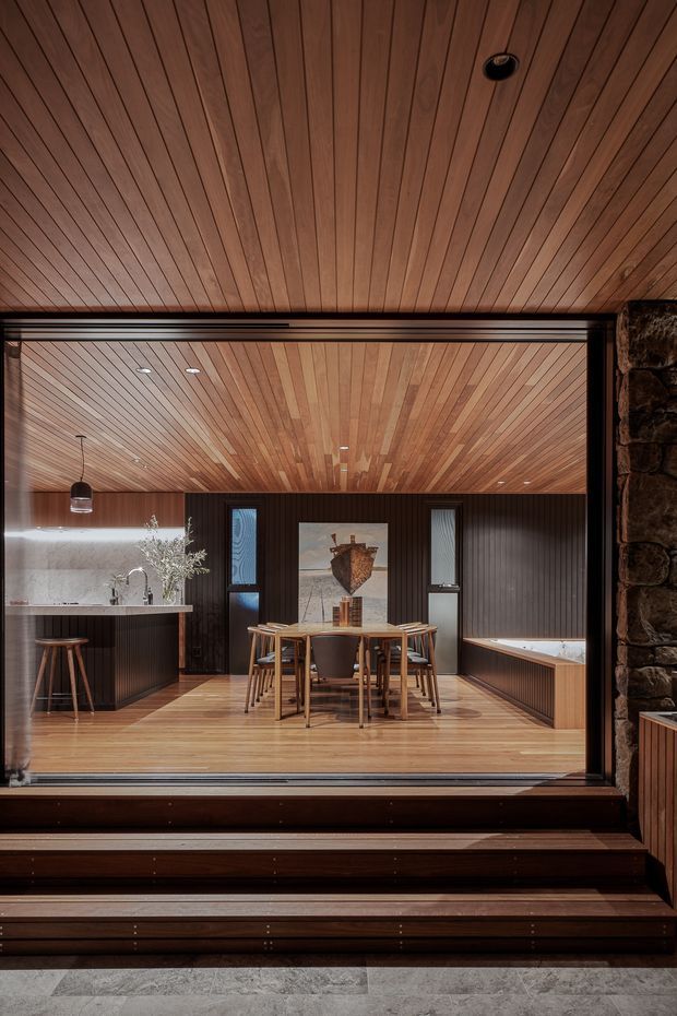 A refined open-plan dining and kitchen interior viewed through a large glazed opening from a stepped threshold. The room features extensive warm timber lining on the ceiling and floor, dark vertical timber wall panelling, a compact kitchen with a pale stone benchtop, a timber dining table with chairs, built-in bench seating, pendant and recessed lighting, and a rugged dark stone wall at the right edge. The composition has a strong indoor-outdoor connection and a warm, contemporary lodge-like atmosphere.