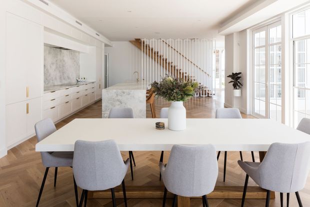 Bright open-plan interior featuring a large dining table in the foreground, a white kitchen with a marble-look island and splashback, and a timber staircase with white vertical balustrades. Full-height glazed doors and windows bring in abundant natural light, while herringbone timber flooring and light upholstered dining chairs create a refined contemporary setting.