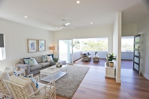 Bright, airy open-plan living room with white vertical wall panelling, pale upholstered seating, a white coffee table, timber flooring, recessed lighting, and a ceiling fan. Large sliding glass doors open onto a furnished balcony or terrace, with additional glazing and an open doorway visible at right. The styling is relaxed coastal with blue-green accents and abundant daylight.