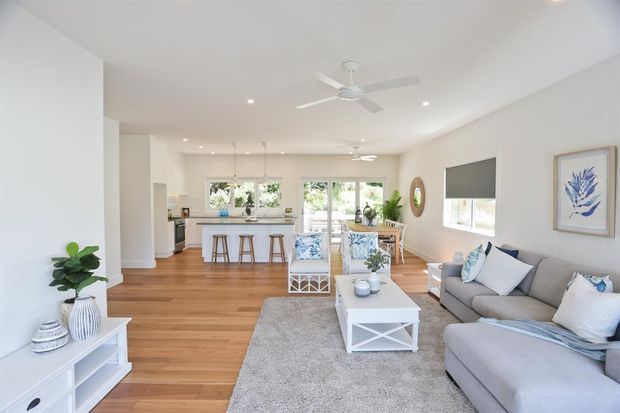 Bright open-plan living room with a grey sectional sofa, white coffee table, light rug, and coastal-inspired decor. The room opens directly into a kitchen and dining area, with warm timber flooring, white walls, vertical wall panelling, large windows, and a white ceiling fan. Natural light creates an airy, relaxed interior.