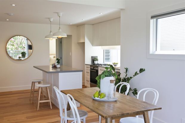Bright open-plan kitchen and dining area with light cabinetry, a compact island with stools, timber flooring, and a rustic timber dining table surrounded by white chairs. Natural light enters through the windows, with indoor plants and a round wall mirror adding warmth and visual interest.