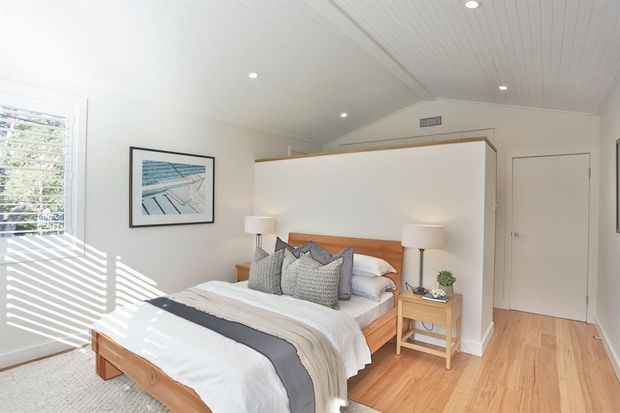 Bright, contemporary coastal-style bedroom with a timber bed and warm timber flooring. The room has white walls, a white V-groove pitched ceiling, plantation shutters, bedside tables with lamps, layered neutral bedding, and framed artwork. A partial-height partition behind the bed separates the sleeping area from the entry or adjacent space.