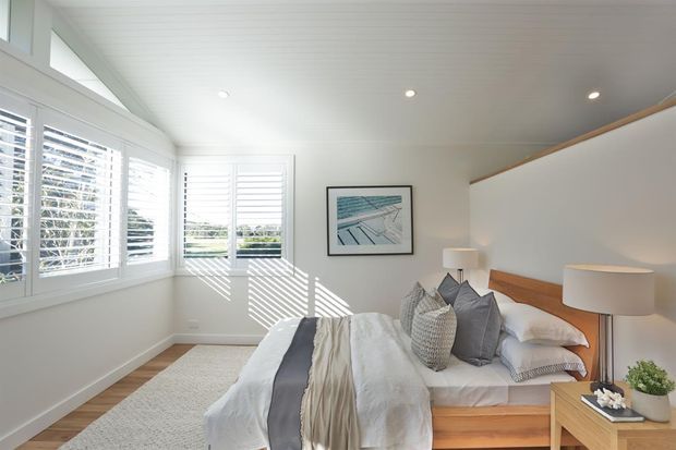 Bright, serene bedroom with extensive white plantation-shuttered windows, a low sloped ceiling with recessed downlights, white walls, and warm timber built-in bed framing and bedside furniture. The bed is dressed in layered neutral bedding with grey and textured cushions, complemented by a pale rug, simple lamps, and a framed coastal artwork.