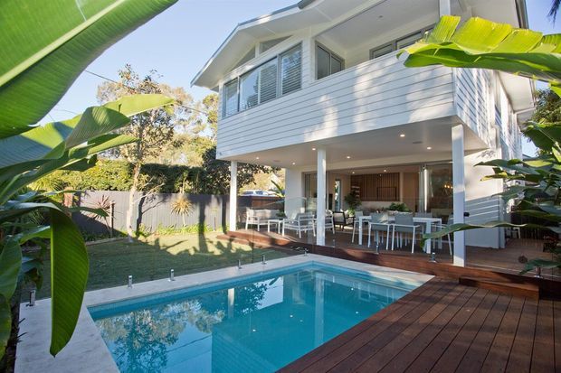 Bright exterior view of a raised, two-storey white-clad house with an open ground-floor entertaining area, dining furniture, timber decking, landscaped garden, and a rectangular swimming pool framed by large tropical banana leaves.