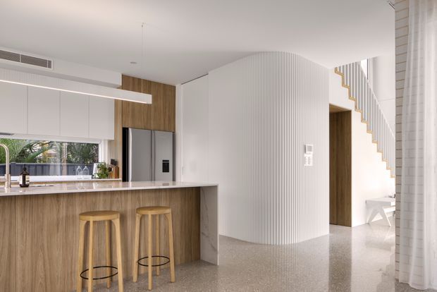 Bright contemporary open-plan kitchen with a timber island and two stools, white cabinetry, stainless-steel refrigerator, pale stone benchtop, and a curved vertically ribbed feature wall. A sculptural white staircase and open living area are visible beyond the kitchen. The space has a clean, refined aesthetic with terrazzo-style flooring and integrated linear lighting.