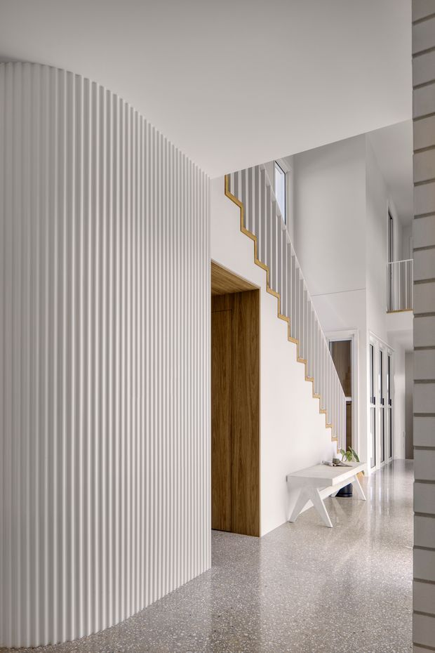 A bright, refined residential hallway and stairway interior. A curved wall clad in closely spaced vertical white ribbing dominates the foreground, leading toward a sculptural staircase with white balustrades and warm timber edging. A tall timber-lined doorway, terrazzo flooring, white walls, internal glazing, and a minimalist console table create a clean contemporary composition.
