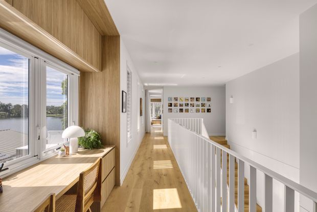 Bright contemporary upper-level hallway or landing with a timber-lined built-in desk and cabinetry beside large windows overlooking water. A white balustrade borders the stair void, with pale timber flooring, white walls, recessed lighting, and a gallery wall at the far end. The space feels airy, warm, and refined.