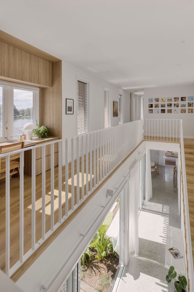 Bright, airy interior view of a contemporary multi-level home showing an upper-floor landing and stair void with white vertical balustrades, pale timber flooring, and white walls. A light timber built-in window seat and cabinetry frame glazing on the left, with a dining area beyond. The stairwell overlooks a lower hallway with extensive glazing, indoor planting, and strong natural light; a gallery wall of small artworks decorates the far landing.