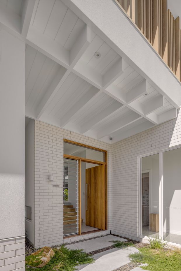 A bright contemporary residential entry courtyard viewed from outside. The facade is clad in white painted brick, with a tall timber front door and matching timber-framed glazing. A white exposed-beam soffit creates a covered entry, while vertical timber battens appear at the upper level. Concrete stepping slabs, gravel strips, and ornamental grasses form the landscaped approach. The open doorway reveals a stairway and part of the interior beyond.