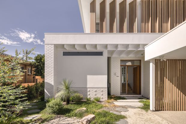 A contemporary residential exterior viewed from the front garden. The white-rendered and white brick facade features strong horizontal and vertical forms, deep roof overhangs, exposed soffit beams, tall upper-level openings screened with vertical timber battens, and a sheltered timber entry door. A timber slatted carport or garage enclosure is visible at right, while native-style planting and stepping stones form the landscaped approach.