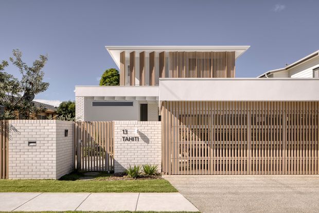 Front exterior view of a contemporary multi-level residence with crisp white rendered and masonry walls, strong horizontal roof planes, and extensive vertical timber screening. A slatted timber garage door and matching entry gate create privacy while allowing filtered light. The property fronts a driveway and paved footpath, with the address marker '13 TAHITI' visible on the boundary wall.