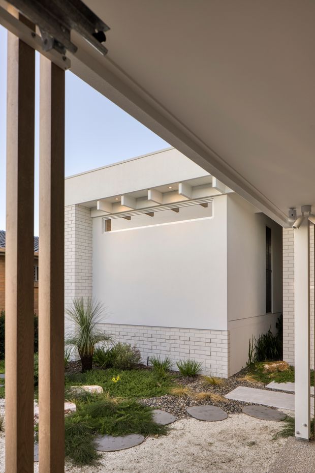 View from a covered outdoor area toward a compact contemporary courtyard. The scene features a white rendered and white brick-clad building, a projecting flat roof or canopy with exposed structural elements, timber posts, and a landscaped gravel garden with stepping stones and grasses. A large sliding or folding door track is visible overhead, suggesting the camera is positioned beside an open glazed enclosure.