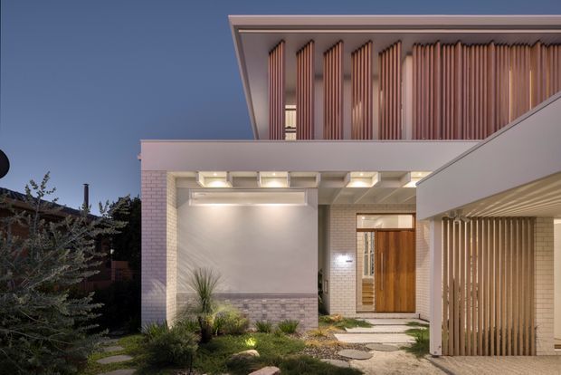 Twilight exterior view of a contemporary multi-level residence. The facade combines pale brickwork, smooth rendered surfaces, warm timber entry doors, and prominent vertical timber screening. Recessed lighting illuminates the covered entry and landscaped front path, while a projecting upper volume and carport-like structure frame the composition.