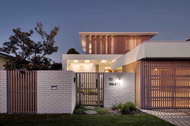 Twilight view of a contemporary multi-level residence with a crisp white rendered facade, projecting flat rooflines, and extensive vertical timber screening. A white brick boundary wall and gated entry display the address, while warm interior and exterior lighting illuminates the recessed entry and slatted garage frontage.