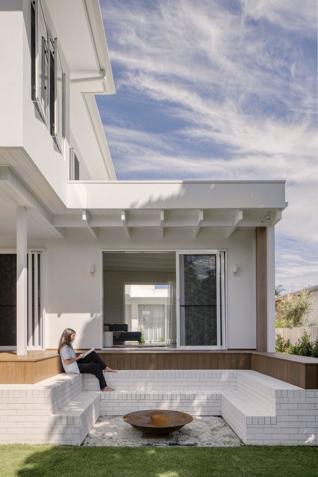 Exterior view of a bright white contemporary home with a flat-roofed covered terrace opening onto a sunken outdoor lounge. The terrace includes built-in white brick bench seating, timber-toned raised planters or decking, a shallow circular fire bowl, and lawn. Large sliding glazing connects the outdoor area to the interior living space, while projecting roof beams and clean geometric forms define the facade.