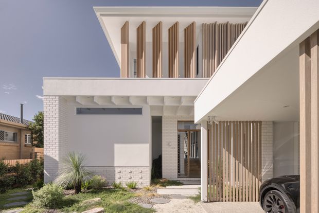 Contemporary two-storey residential facade viewed from the front, featuring white brickwork, crisp rendered balcony and roof forms, extensive vertical timber screening, and an integrated carport. A landscaped entry path and drought-tolerant planting sit in front of the home, with neighbouring suburban housing visible at the sides.