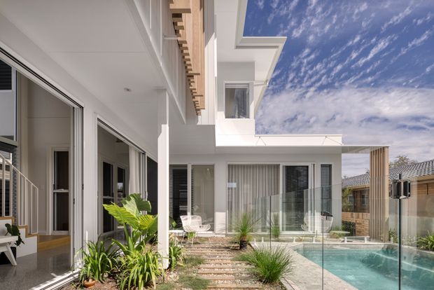 Bright contemporary multi-level residential courtyard viewed from an outdoor garden area, with white rendered walls, extensive glazing and sliding doors, timber architectural screening, a glass pool fence, and a compact turquoise swimming pool. Tropical planting and stepping-stone paving soften the crisp geometry, while a dramatic open-to-sky void frames the blue sky.