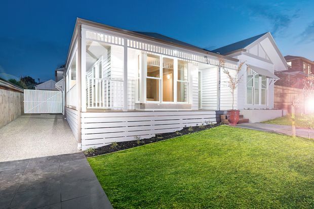 Exterior view of a bright white single-storey house with a pitched gable roof, horizontal weatherboard cladding, enclosed front verandah, decorative timber screens and white-framed windows. A paved path, driveway, front lawn and neighbouring homes are visible, photographed at dusk with strong exterior lighting.