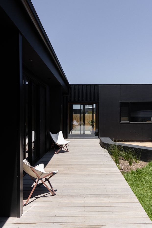 A contemporary black-clad single-storey house arranged around a timber deck walkway or courtyard. Full-height dark-framed glazing lines the left facade, while a low dark wing with a horizontal window borders the right. Two folding lounge chairs sit on the pale timber decking, with sparse ornamental grasses and lawn beside the building under a clear sky.