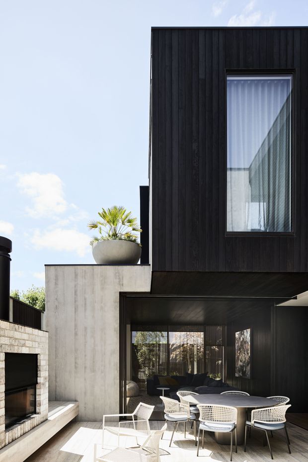 Contemporary multi-level home exterior with a dramatic black vertical-clad upper volume, raw concrete walls, large glazed openings, and a covered outdoor dining terrace. A built-in masonry fireplace, white outdoor dining furniture, and a sculptural planter occupy the sunlit courtyard-like area.