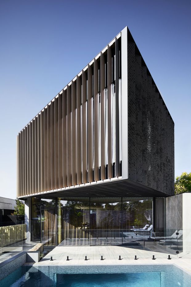 A striking modern multi-level building with a cantilevered upper volume, sharply angled roofline, and a facade screened by repeated vertical fins. Dark perforated cladding forms the side wall, while extensive floor-to-ceiling glazing encloses the lower level. A swimming pool and paved terrace occupy the foreground, with a covered outdoor lounge visible beneath the cantilever.