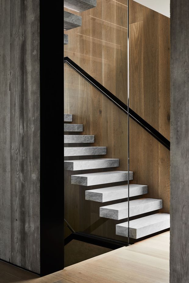 A dramatic contemporary stairway with floating concrete steps rising beside a warm timber-panelled wall. Black metal handrails and framing contrast with dark textured concrete walls, while a clear glass balustrade provides visual separation. Pale timber flooring is visible at the landing.