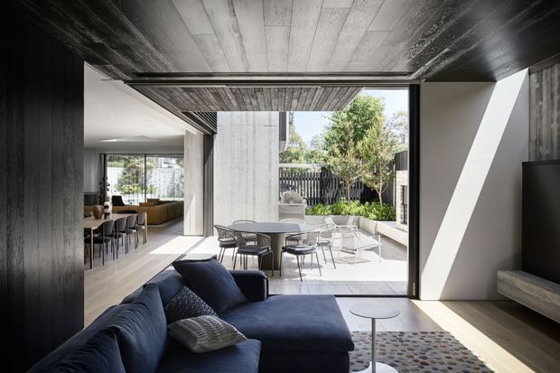 A contemporary open-plan living room with a deep navy sectional sofa in the foreground, opening onto a bright dining area and landscaped courtyard. The space features dark timber wall and ceiling finishes, light timber flooring, strong concrete or plaster architectural forms, expansive glazing, and a covered outdoor dining terrace.