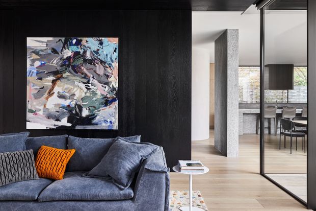 Contemporary open-plan living area with a deep charcoal sofa, patterned cushions, and a bold abstract artwork against a dark timber feature wall. Full-height black-framed glazing separates the living room from a bright dining area, with pale timber flooring, exposed concrete or stone-textured structural elements, and a neutral modern palette.