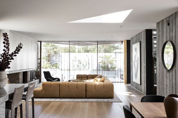 Bright open-plan living and dining area with a warm neutral palette, light timber flooring, a large tan sectional sofa, and floor-to-ceiling glazing opening toward an outdoor setting. The room features a skylight, a dark textured feature wall with a round mirror, built-in cabinetry and fireplace, and a dining table in the foreground.