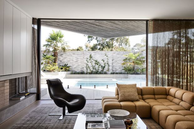 Contemporary living room with a sculptural black lounge chair, large tan sectional sofa, built-in fireplace and cabinetry, and full-height glazing opening onto a covered outdoor terrace and swimming pool. Vertical screened curtains and a textured masonry garden wall reinforce the indoor-outdoor connection.