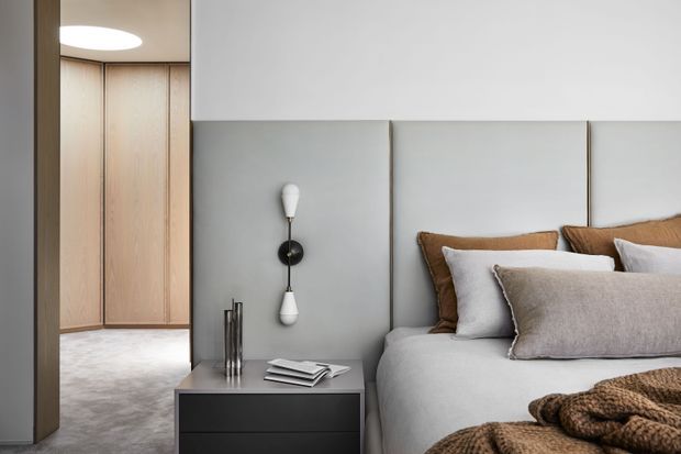 A refined contemporary bedroom with a partially visible built-in timber wardrobe and carpeted dressing area behind the bed. The bed features a tall upholstered grey headboard, layered neutral bedding, brown cushions, and a textured throw. A dark bedside table with sculptural wall lighting and books completes the minimalist composition.