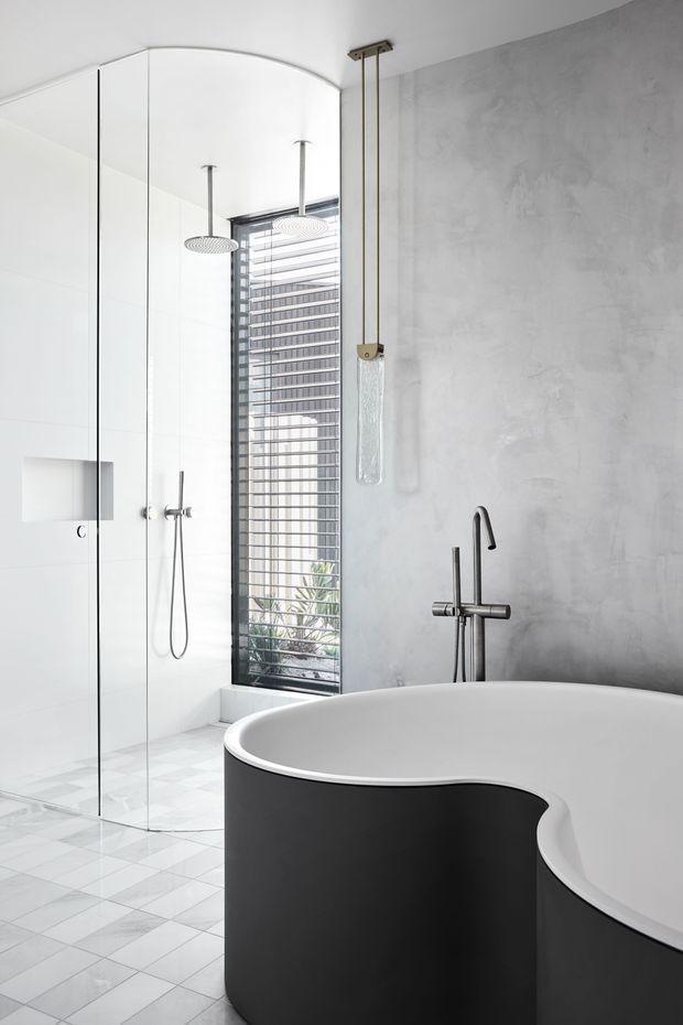 A bright contemporary bathroom featuring a sculptural freestanding black-and-white bathtub, a frameless glass shower enclosure with twin overhead rain showers, a wall-mounted shower set, a grey textured plaster wall, and a tall louvred window. The floor is finished in pale geometric tiles, with minimalist brushed-metal fixtures and a hanging pendant light.