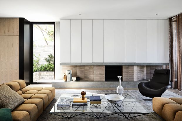 A refined contemporary living room with a low, deep upholstered sectional sofa, sculptural black lounge chair, and glass coffee table over a patterned rug. A full-width built-in media and storage wall combines pale cabinetry, a grey stone ledge, and a textured beige brick feature around a dark fireplace or media recess. Black-framed glazing, timber cabinetry, and full-height curtains complete the room.