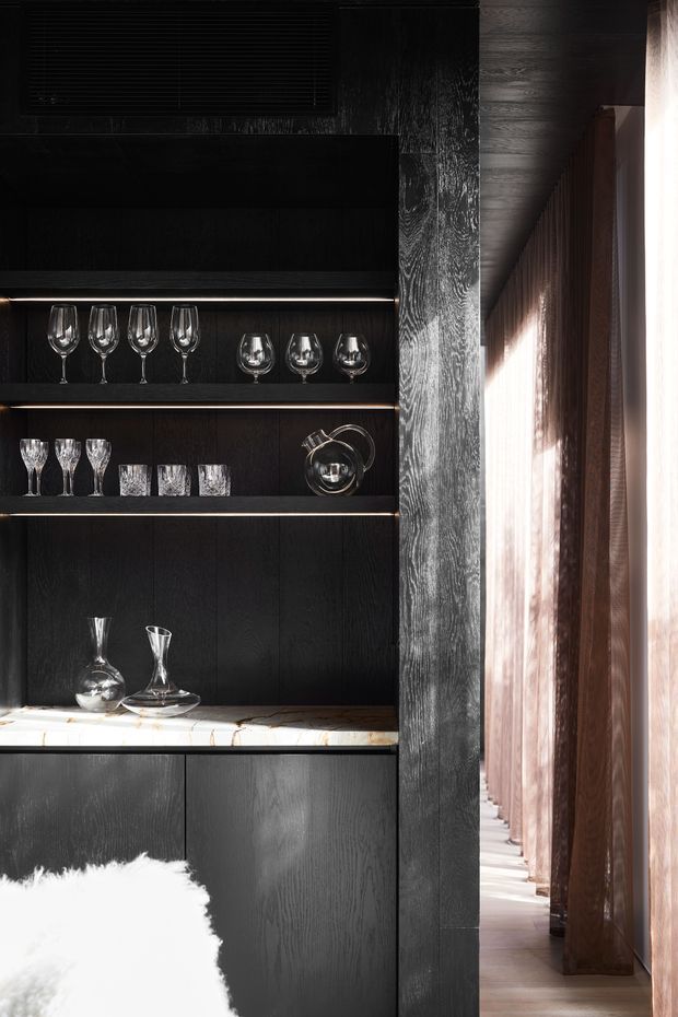 A dramatic, dark built-in display and bar cabinet with open shelves holding wine glasses, tumblers, decanters and a metal vessel. The cabinetry and surrounding walls feature dark stained timber with pronounced grain, integrated linear shelf lighting, and a light stone benchtop. A corridor or glazed opening with warm timber screening and bright daylight is visible to the right. The image has a refined, moody, high-contrast interior aesthetic.