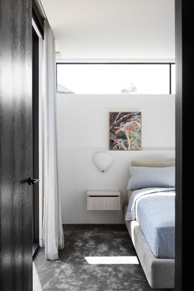 Bright contemporary bedroom viewed through a partially open dark timber door. The room features white vertical wall panelling, a high horizontal window, sheer white curtains, grey carpet, a light upholstered bed with pale blue bedding, a floating bedside table, and abstract artwork. The space has a clean, refined, softly layered interior with strong natural light.