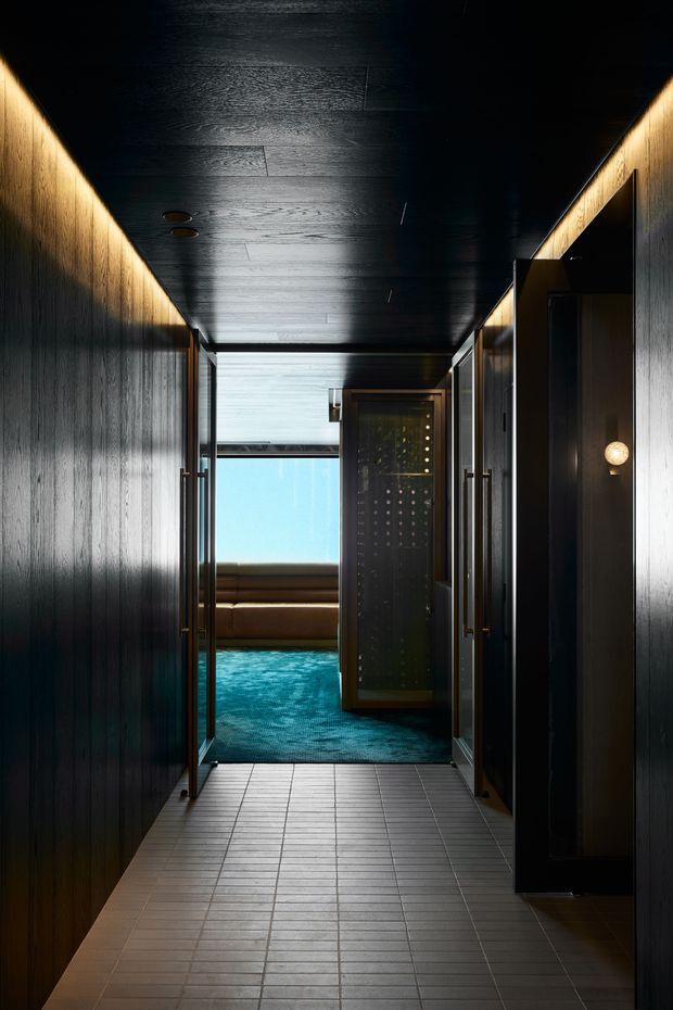 A dark, dramatic interior corridor lined with vertically grained dark timber wall and ceiling panels, recessed lighting, and multiple doorways. The tiled floor leads toward a bright blue-water area visible at the far end, with a glass-fronted enclosure and warm accent lighting creating a luxurious spa-like atmosphere.
