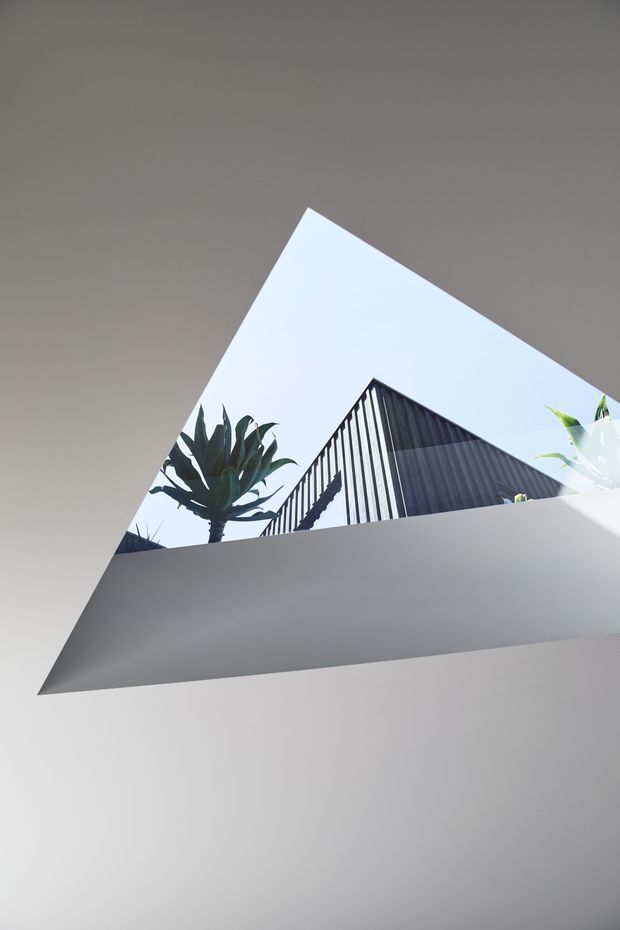 Abstract architectural view looking upward through a sharply angled opening or courtyard. Smooth white rendered surfaces frame a bright sky, with a sculptural tropical plant and a dark vertically ribbed triangular roof or facade visible beyond. The image emphasizes geometric forms, strong diagonals, and a minimalist contemporary composition.
