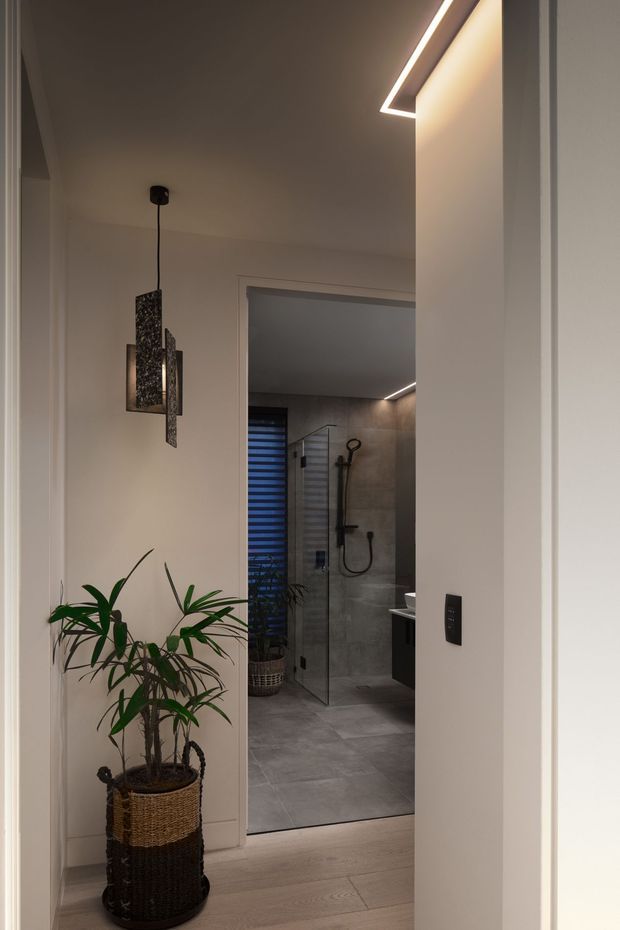 A warmly lit modern hallway leading into a contemporary bathroom. The foreground features pale timber flooring, white walls, a woven planter with a tall green plant, a sculptural dark pendant light, and integrated linear ceiling lighting. Through the open doorway is a grey tiled bathroom with a frameless glass shower, black shower fittings, dark vanity cabinetry, and a narrow window with a privacy blind.