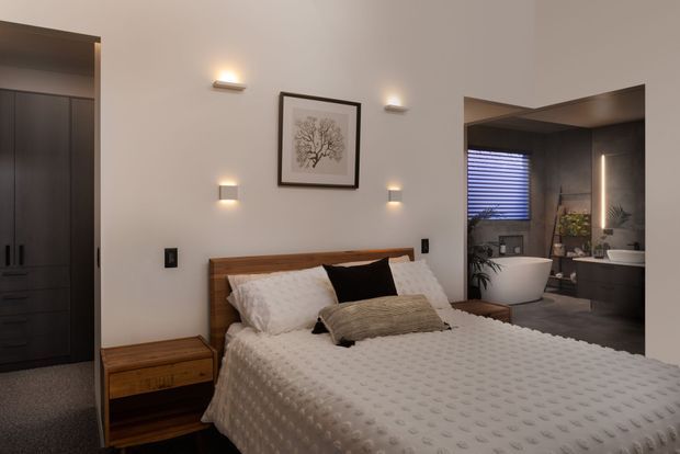 A warm, contemporary bedroom with a wooden bed frame, textured white bedding, bedside table, dark built-in wardrobe, wall-mounted bedside lights, and framed botanical artwork. An open ensuite bathroom is visible to the right, featuring a freestanding bathtub, vanity, dark cabinetry, and a window with blinds.
