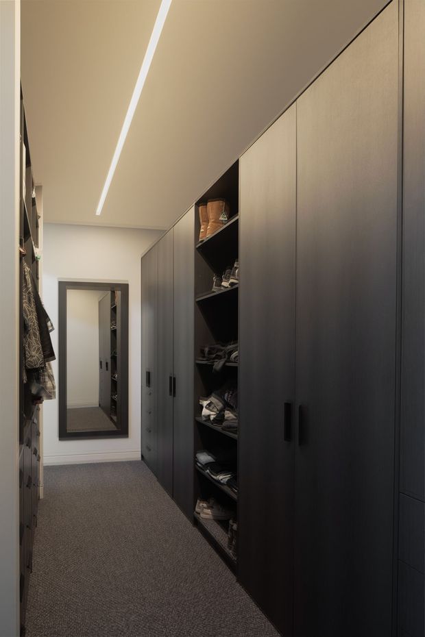 A narrow walk-in wardrobe with floor-to-ceiling dark built-in cabinetry, open shelving displaying folded clothing and footwear, hanging garments along the left side, and a full-length wall mirror at the end. The space has grey carpet, white walls, and a minimalist linear ceiling light.