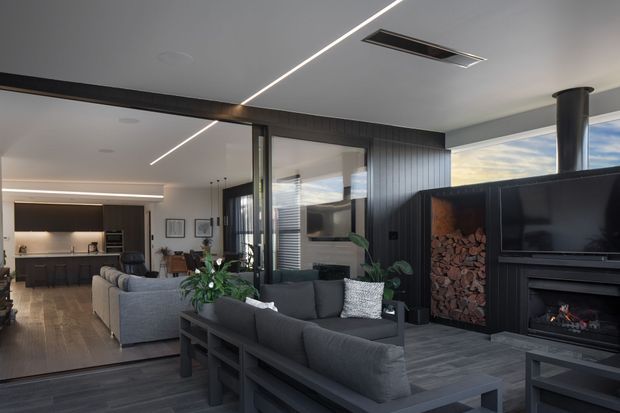 Contemporary open-plan living room with dark timber-look wall panelling, charcoal sofas, a large television and built-in fireplace with stacked firewood. The space opens to a kitchen and dining area, with extensive glazing, linear ceiling lighting and a clean, refined fit-out.